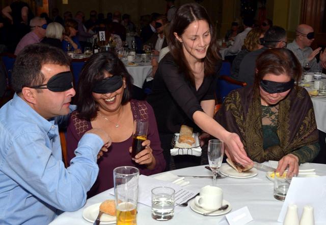 Waiting staff serve diners sat at a table wearing blindfolds 