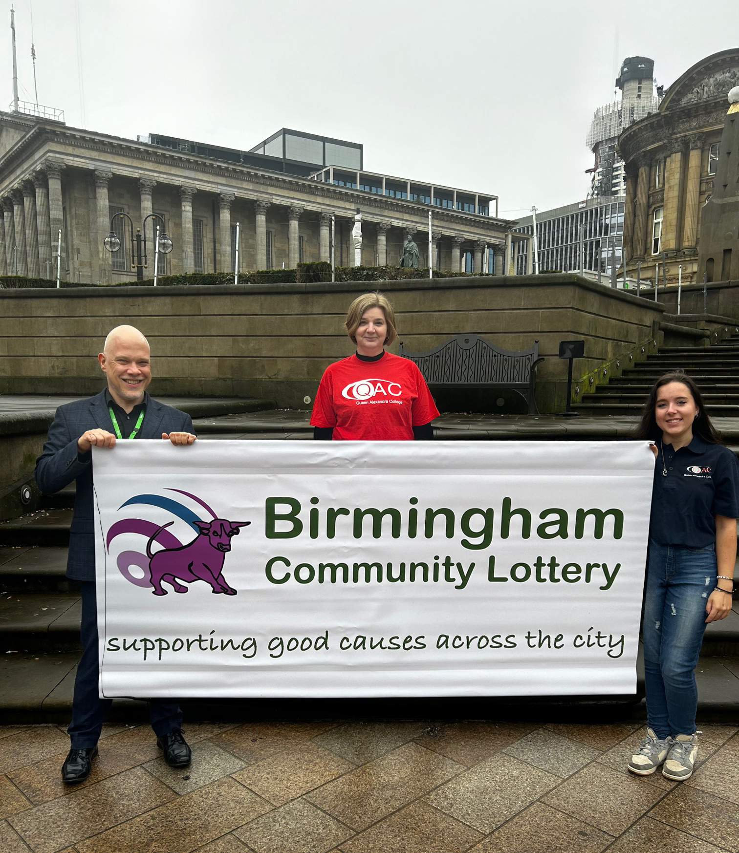 Two QAC members of staff and BVSC Chief Executive smiling with a Lottery banner