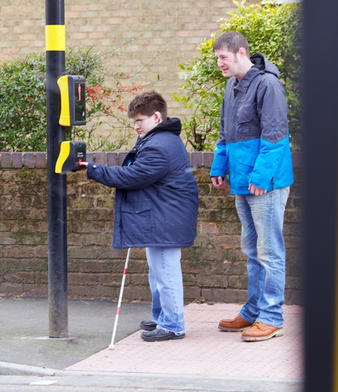 Mobility Officer and a young man using a long cane stand at a pedestrian crossing waiting to cross the road